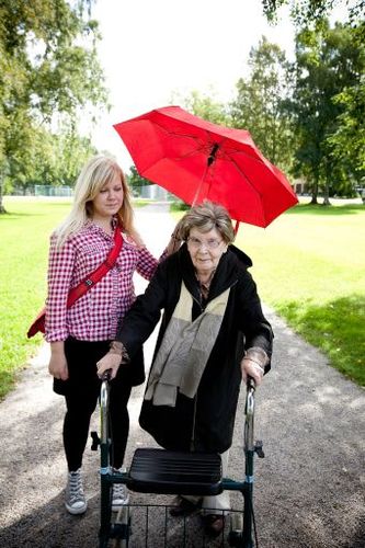 En ung kvinna håller ett paraply över en äldre dam med rollator.